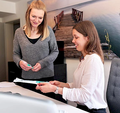 Zwei junge Frauen, Mitarbeiterinnen im Kompetenzzentrum Blase und Darm der Hohenzollern Apotheke schauen sich an einem Schreibtisch Fertigspritzen zur Instillation in die Blase an. Das Bild ist von leicht unten fotografiert.